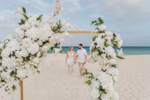 Couple walks hand-in-hand under a white floral arch on a pristine beach during a romantic destination wedding ceremony.
