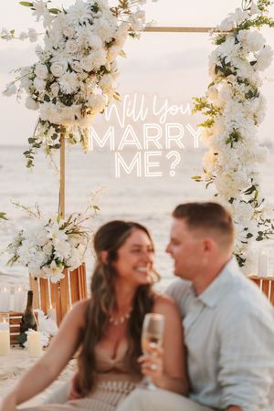 Couple smiling at beach proposal setup with white floral arch and Will You Marry Me neon sign.
