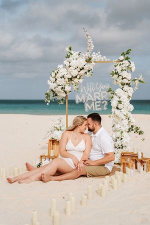 Couple sits on beach sand under floral arch with Will You Marry Me sign, surrounded by candles at romantic proposal setup.