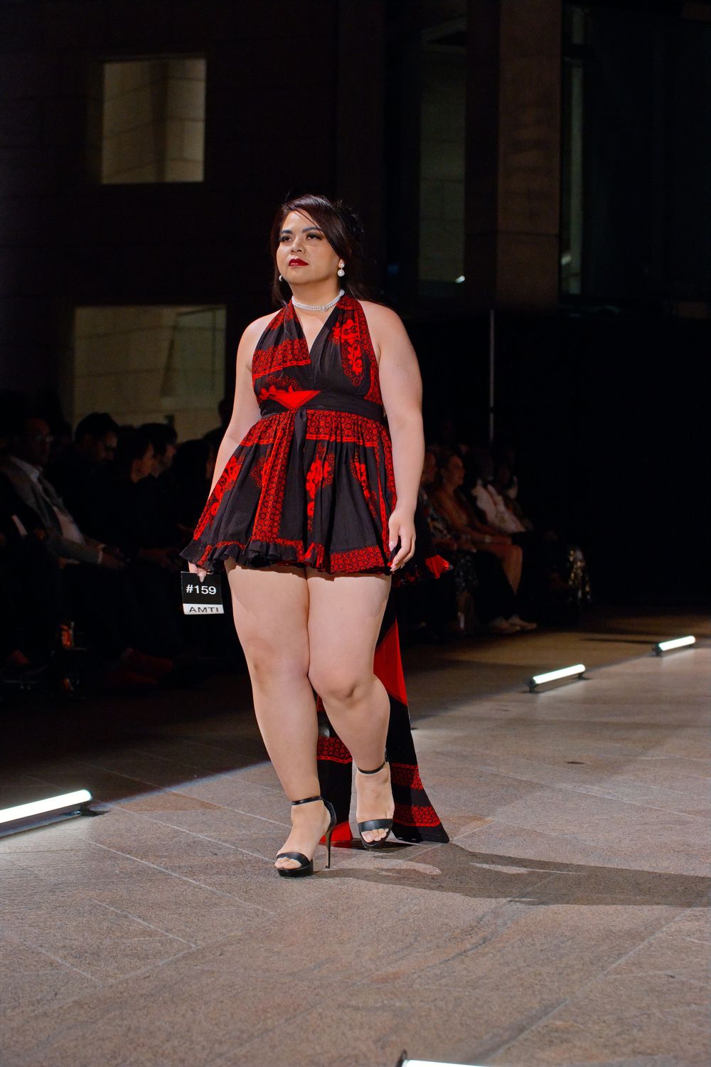 Plus-size model walks runway in red and black plaid mini dress with halter neckline at fashion show.