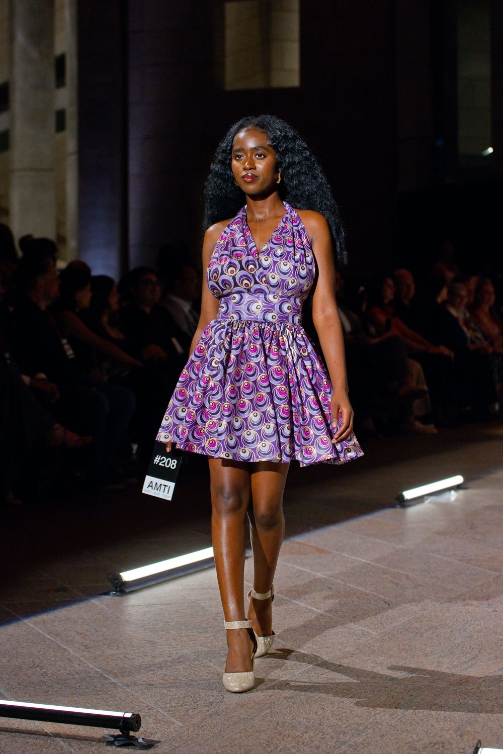 Model walks runway in vibrant purple African print halter mini dress at fashion show.