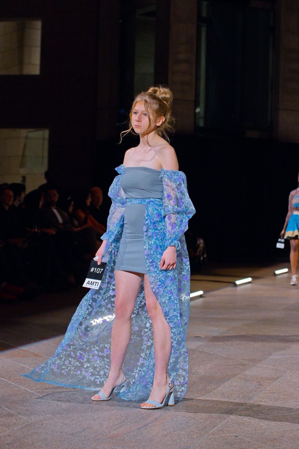 Model walks runway in blue off-shoulder mini dress with sheer floral overlay coat.
