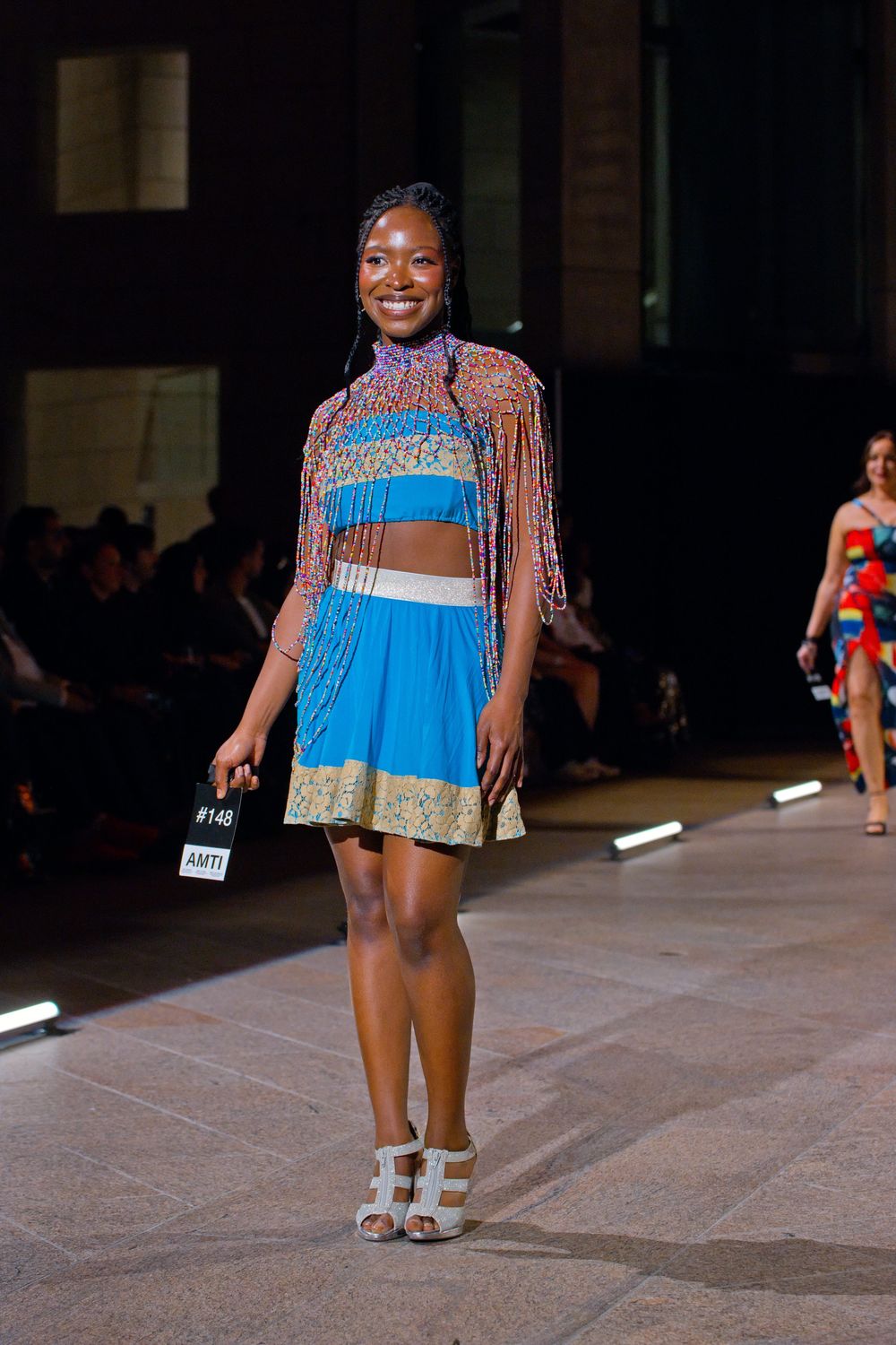 Model walks runway in vibrant blue mini skirt and colorful fringed crop top at fashion show.