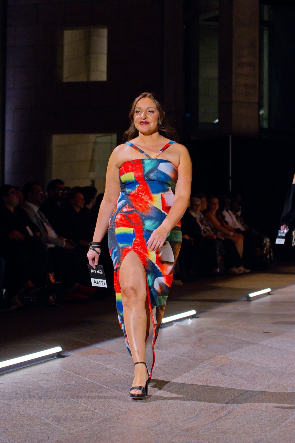 Curvy model walks runway in colorful geometric print dress with red feather detail and black strappy heels.