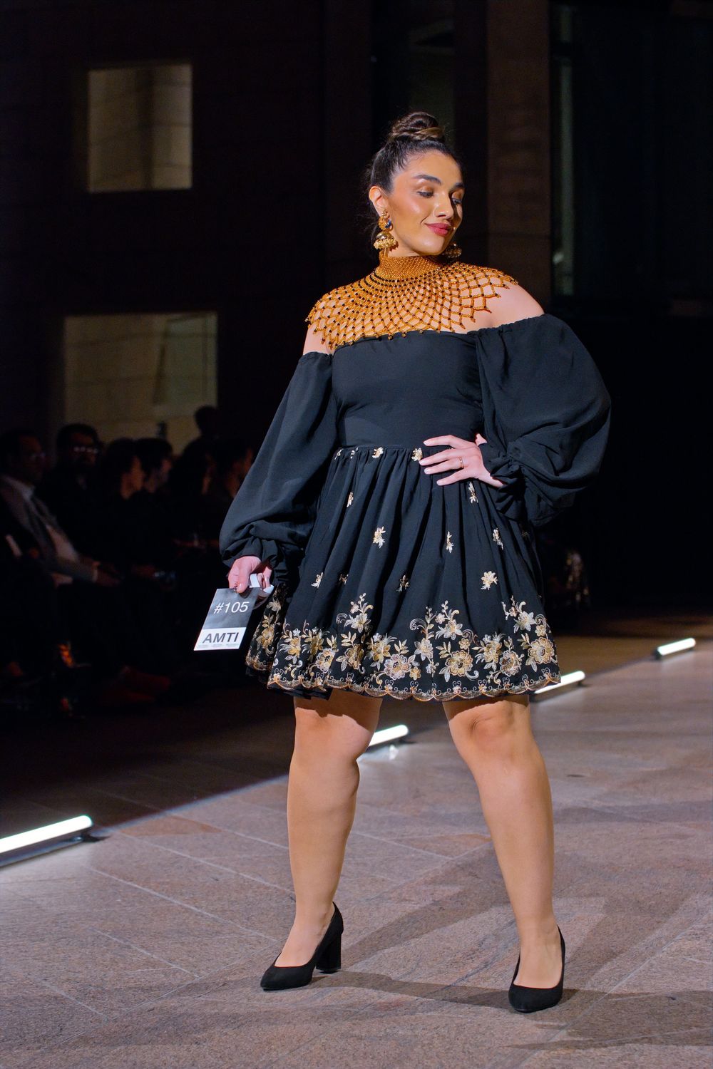 Model walks runway in black floral mini dress with gold neckpiece and dramatic sleeves at fashion show.