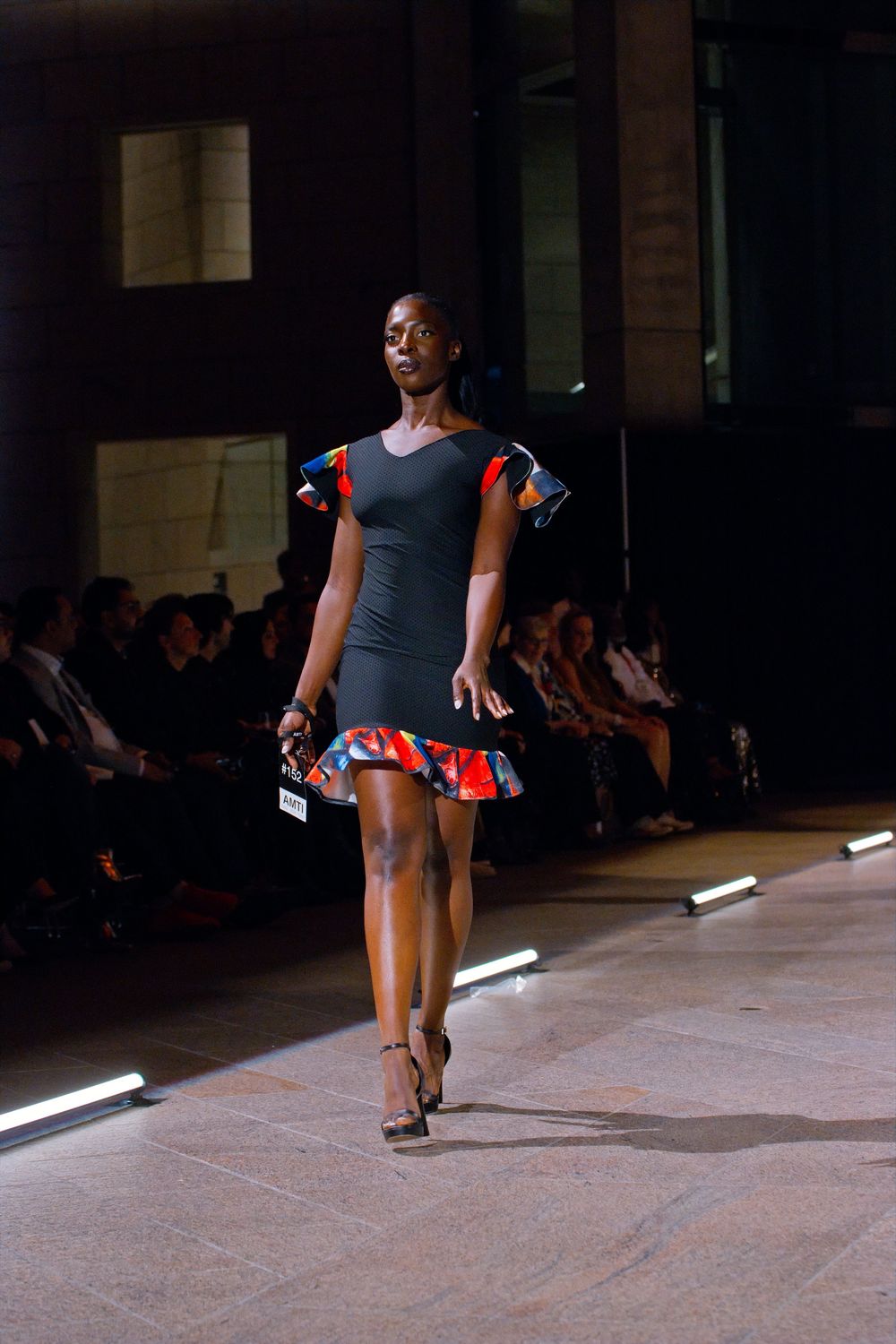 Model walks runway in black dress with colorful ruffled sleeves and hem at fashion show.
