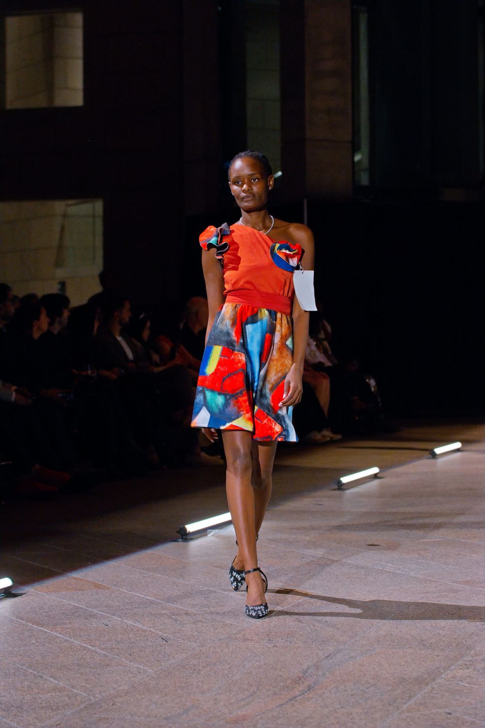 Model walks runway wearing vibrant red one-shoulder dress with colorful abstract print skirt at fashion show.
