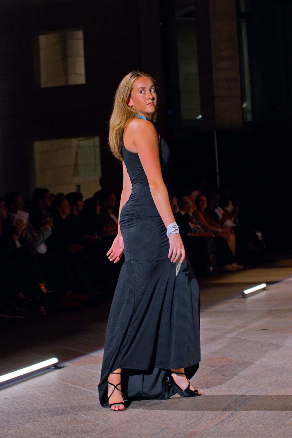Blonde model walks fashion runway in elegant dark gray sleeveless mermaid gown with strappy black heels.