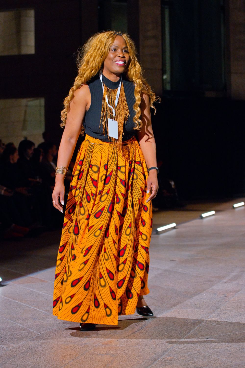 Model walks runway wearing vibrant orange African print maxi skirt with black sleeveless top.
