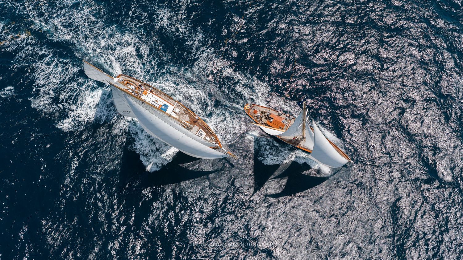 Aerial view of two elegant sailing yachts racing side by side on deep blue ocean waters, leaving white wake trails.