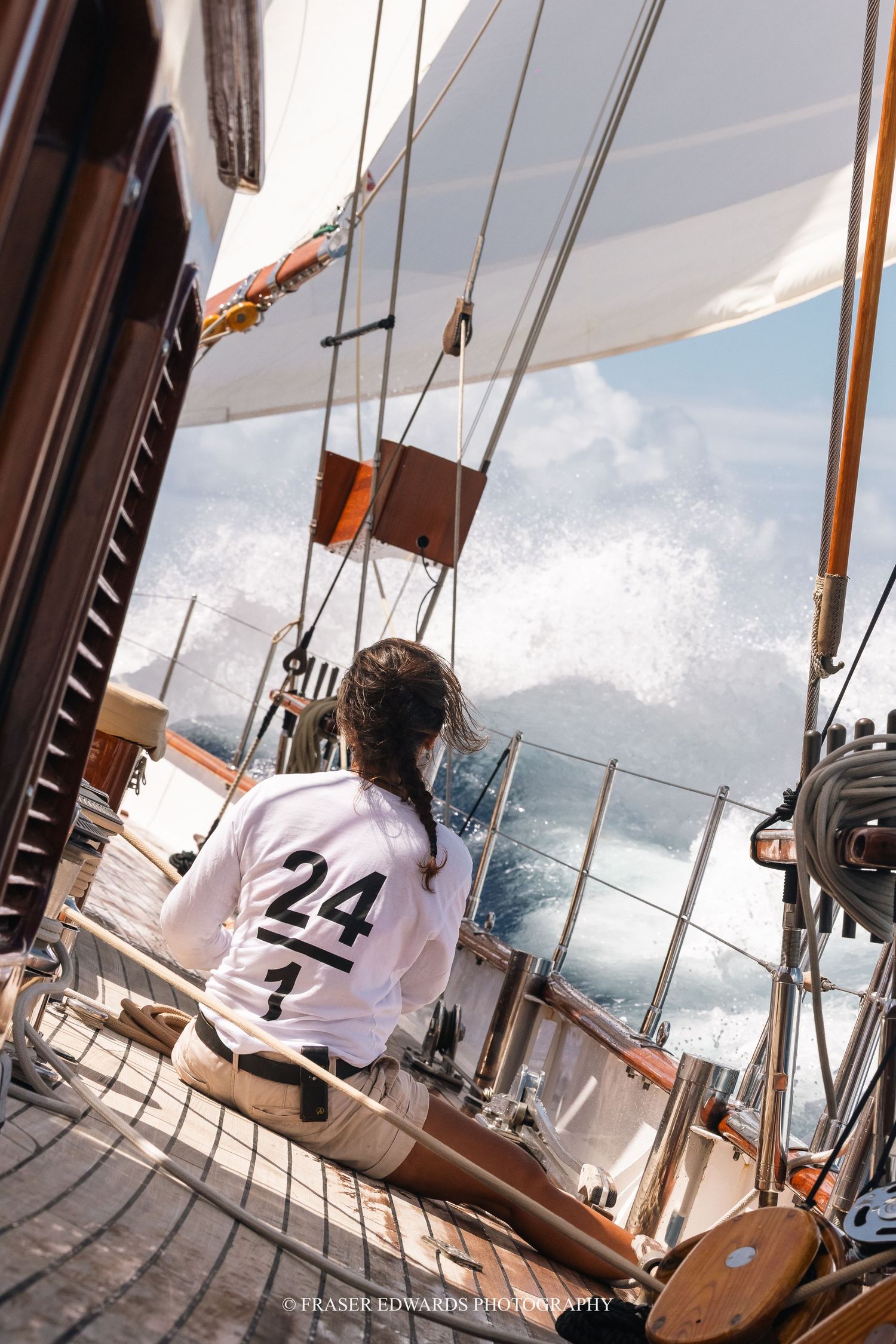 Sailor in white racing shirt number 24 sits on classic wooden sailboat deck watching waves during regatta race.