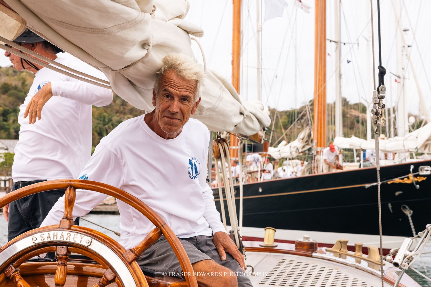 Experienced sailor at the helm of a classic wooden sailboat in a marina, wearing a white shirt.