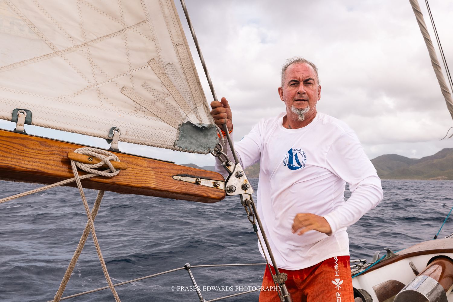 A sailor in a white long-sleeve shirt stands on a classic wooden sailboat deck with mountains visible in the background.