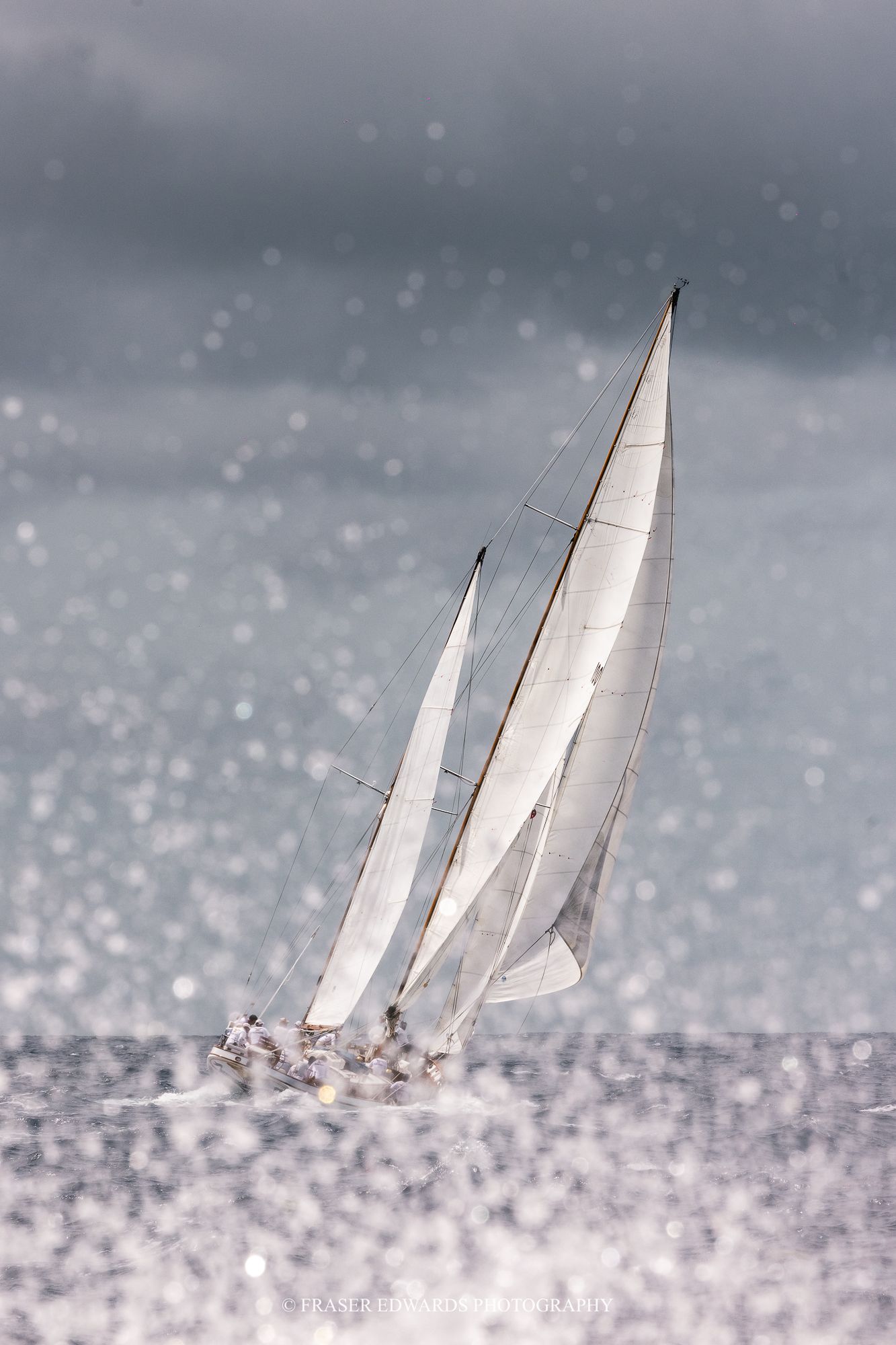 Two white-sailed yachts race through sparkling sunlit ocean waters under dramatic stormy grey skies.