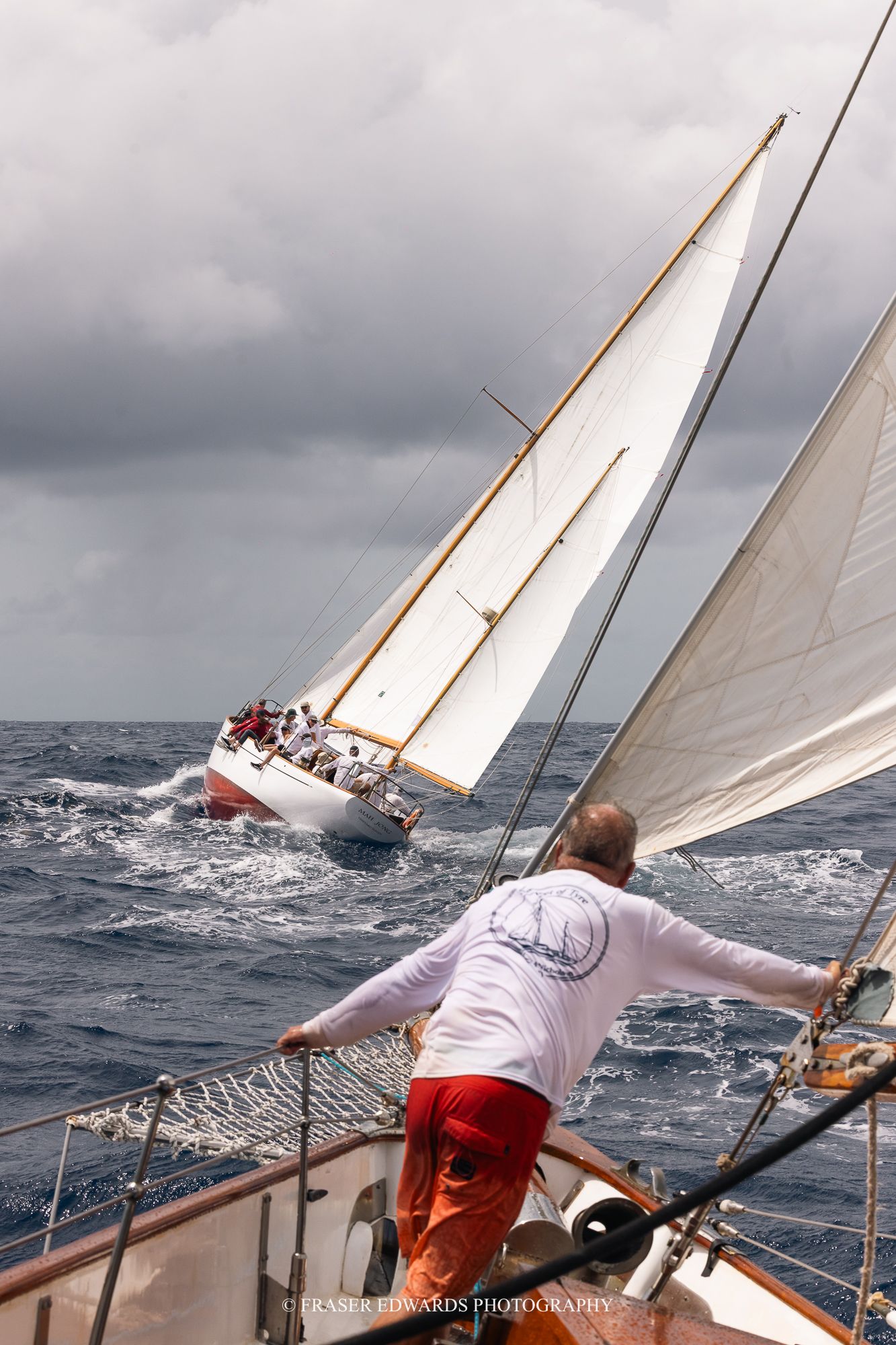 Sailor in red pants watches a classic wooden sailboat racing through choppy seas under stormy skies.