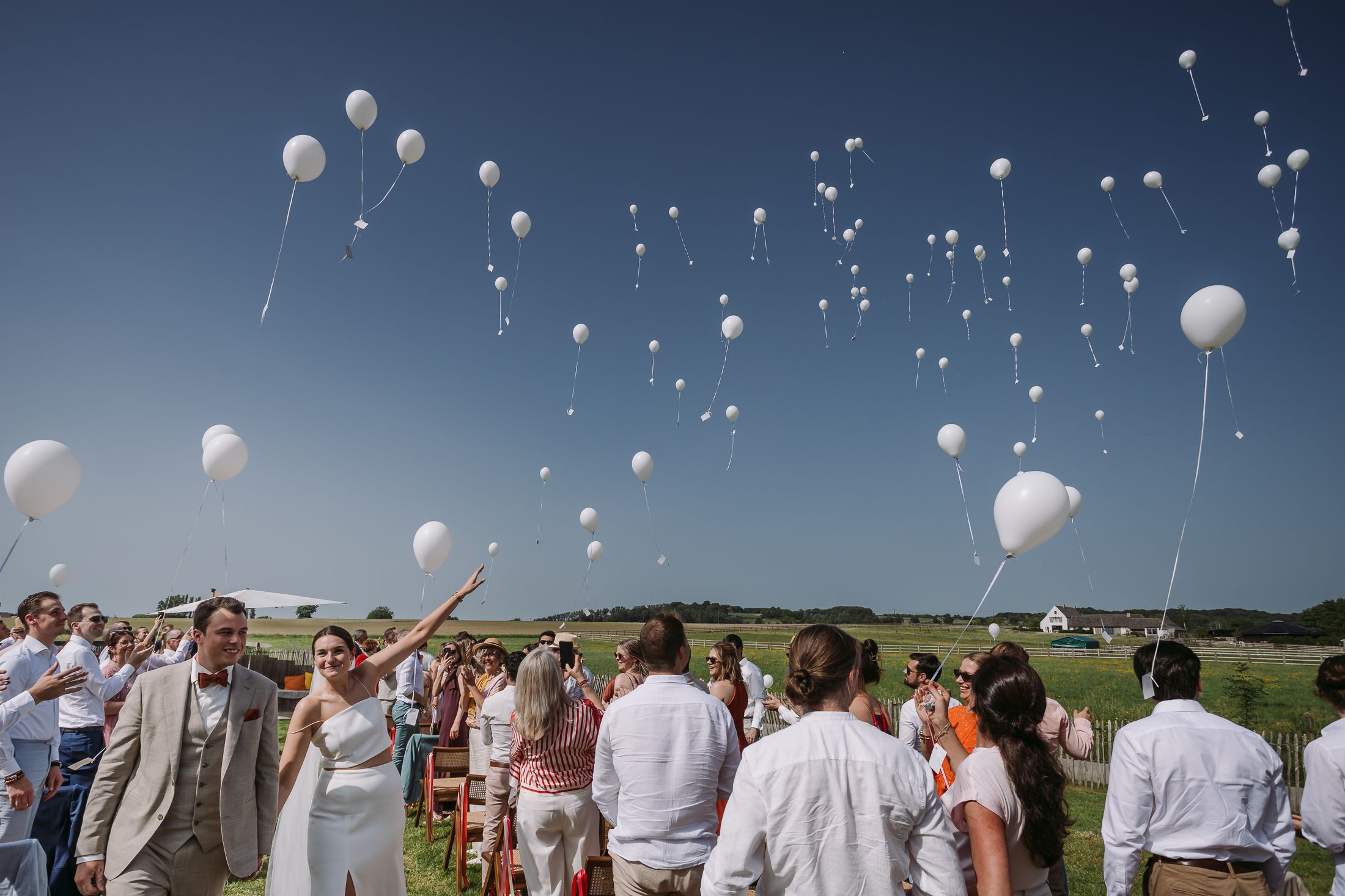 Wedding guests in white release white balloons into a clear blue sky at an outdoor celebration.