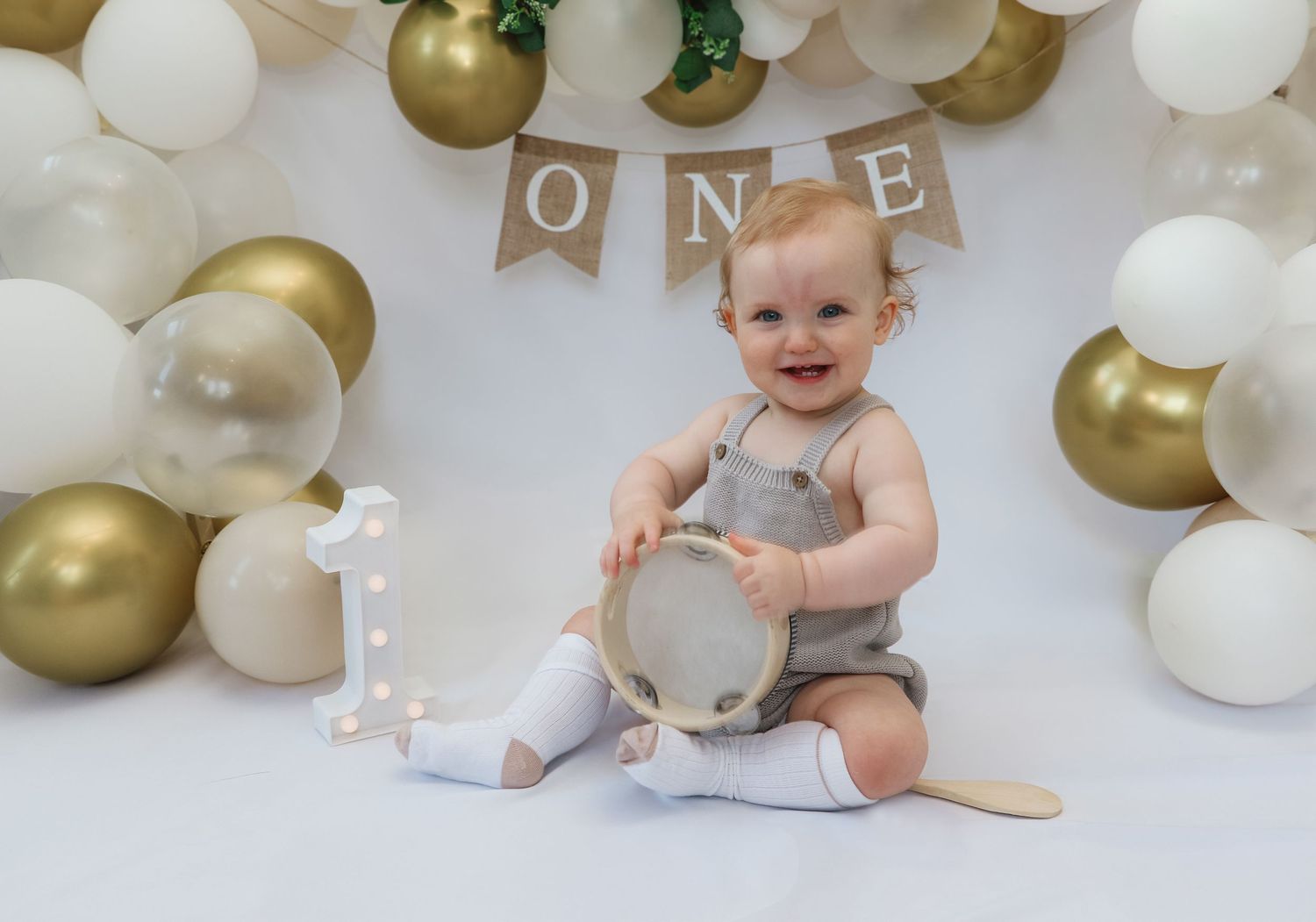 Baby celebrates first birthday with gold and white balloons, holding a tambourine in front of an ONE banner.