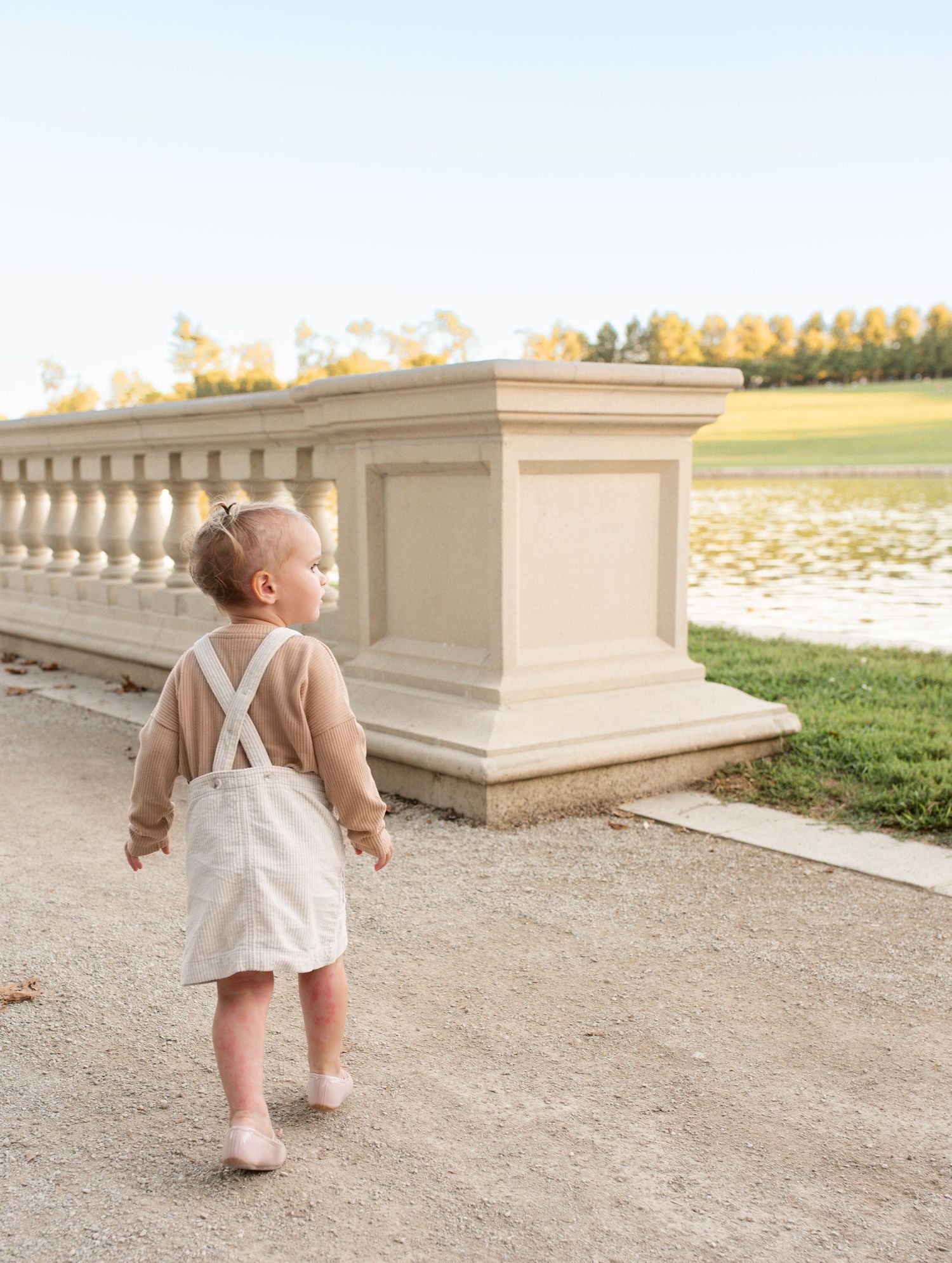 Toddler in white overalls and tan sweater walking toward ornate stone balustrade near grand basin at forest park