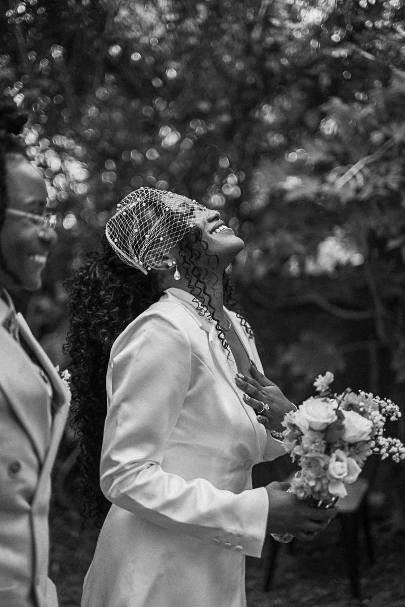 Black and white photo of a joyful bride in white suit and veil headpiece holding bouquet, laughing outdoors.