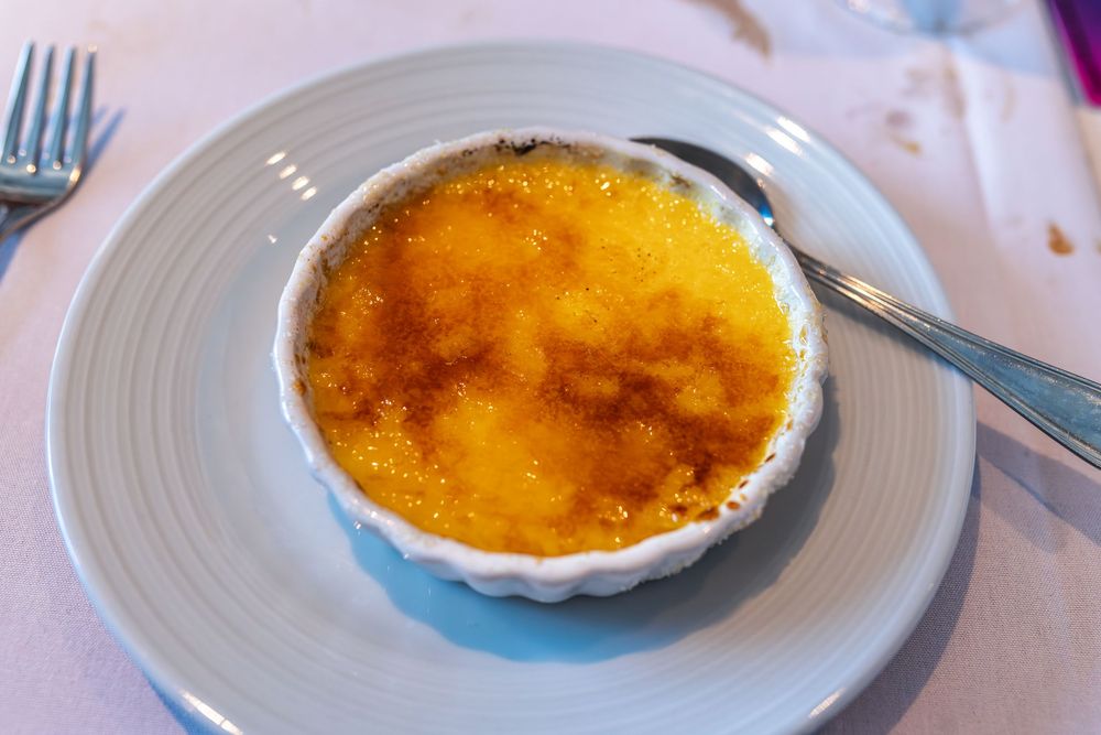Golden-brown crème brûlée in a white ramekin on a gray plate, served with a spoon in an elegant restaurant.