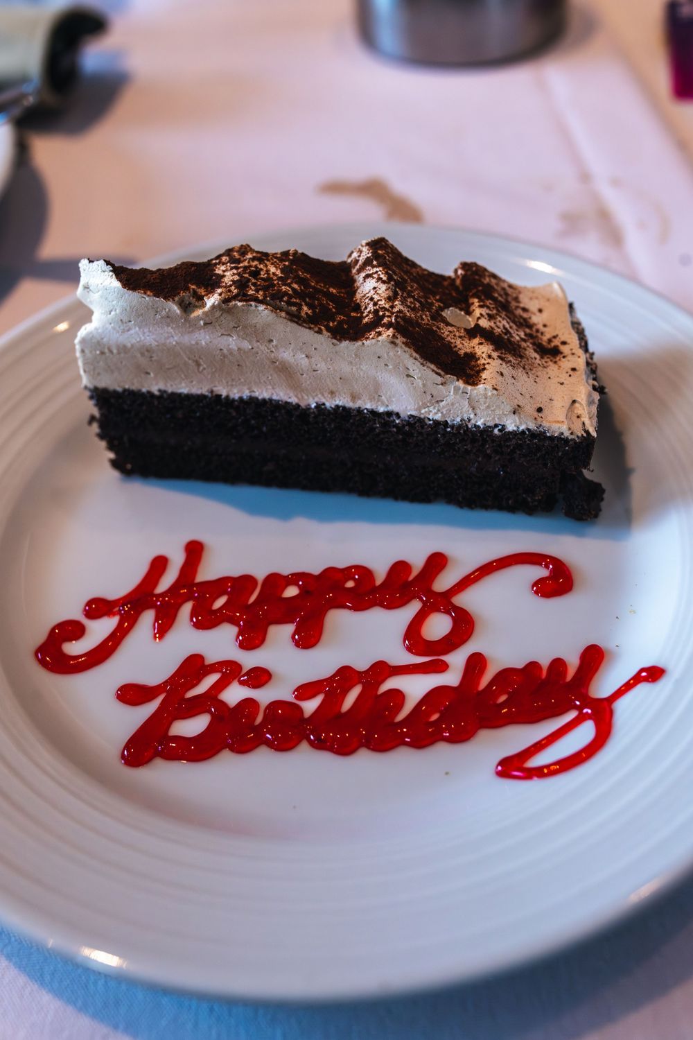 A slice of chocolate birthday cake with whipped cream topping on a white plate with red Happy Birthday syrup writing.