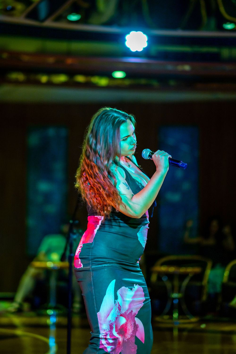 A female singer performs on stage holding a microphone, wearing a black floral dress under colorful stage lighting.