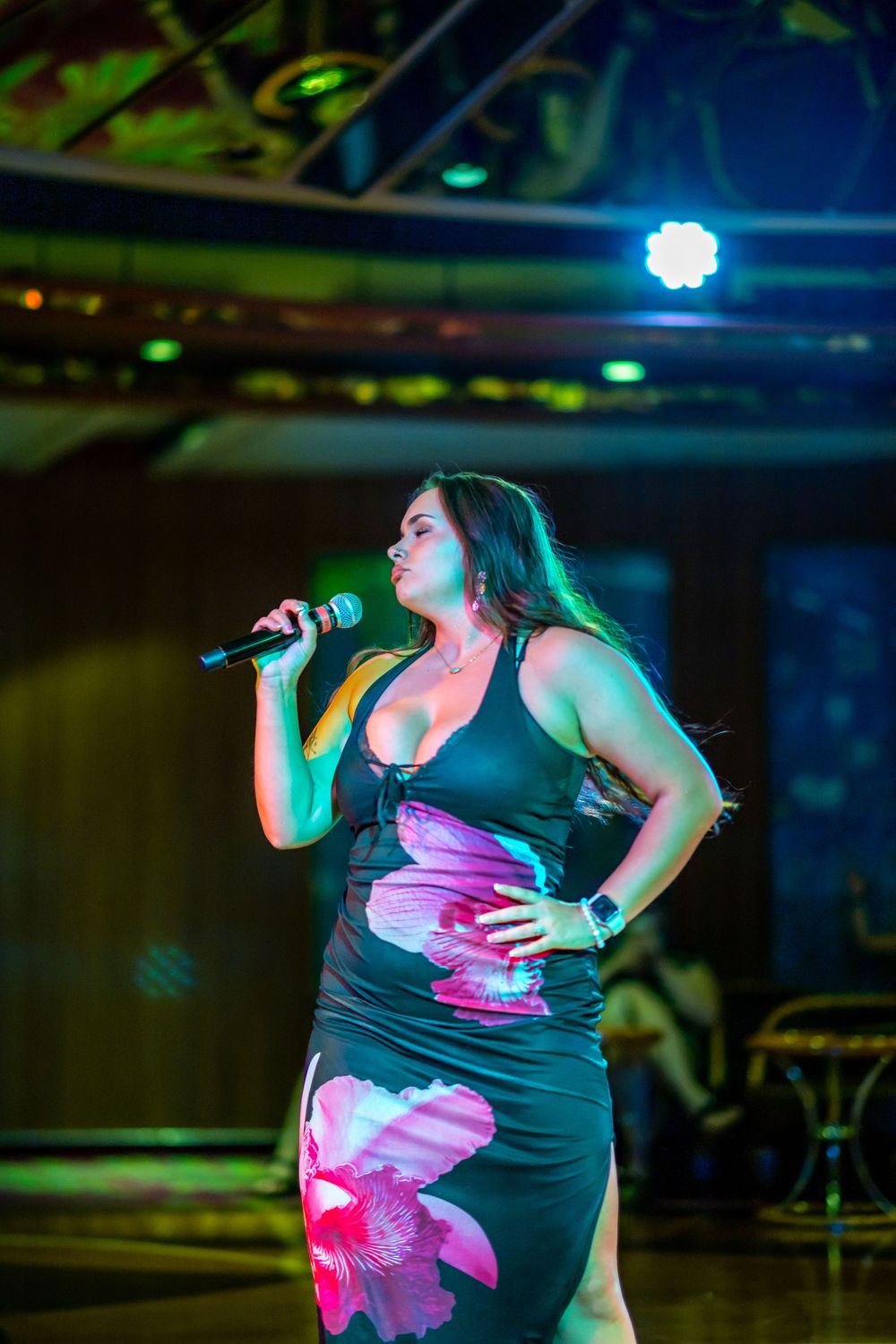 Woman in floral dress singing on stage with microphone under colorful stage lights.