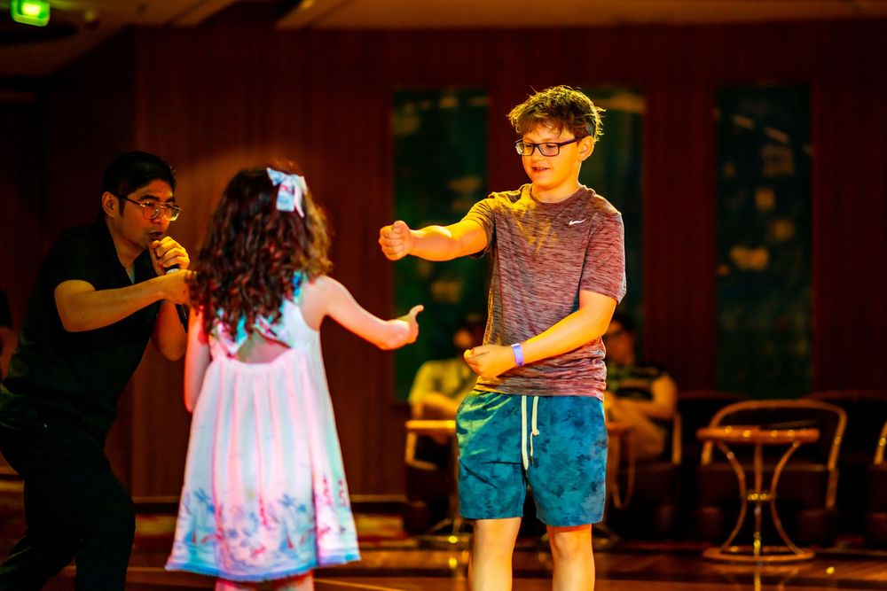 Two teens dance together on a stage while a man watches in a warmly lit ballroom setting.