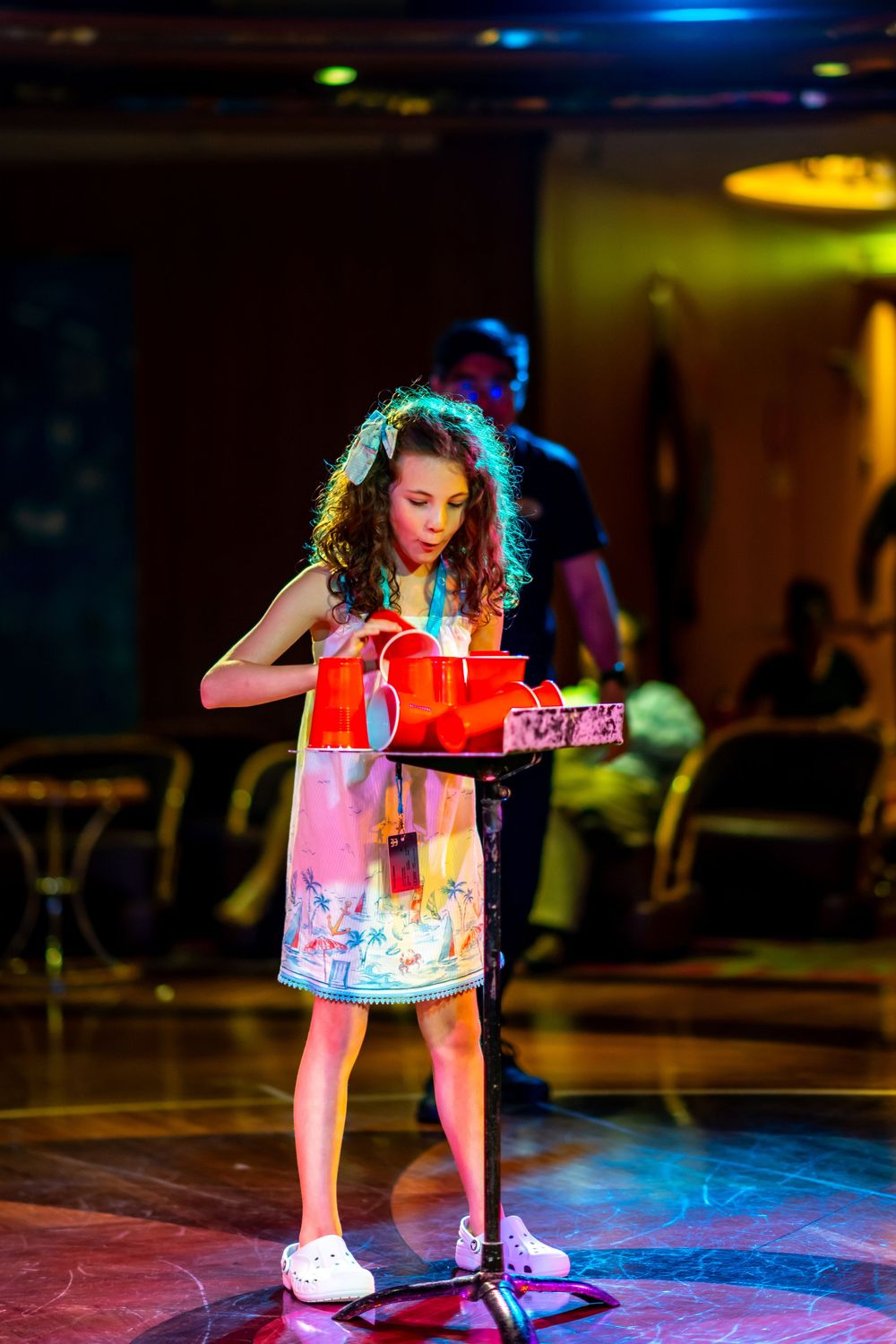 A curly-haired girl in a floral dress performs on stage with red props under colorful lights.