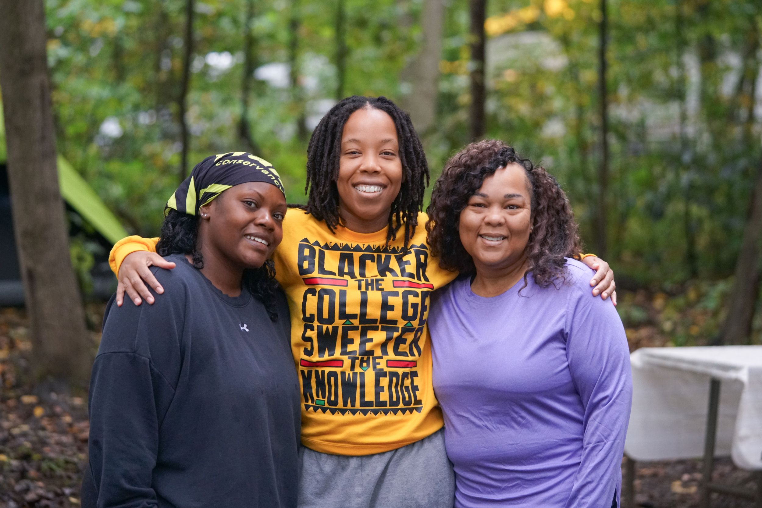 Three women smiling outdoors, center wearing a Blacker the College Sweeter the Knowledge shirt, posing together in wooded area.