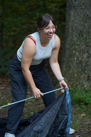 Smiling woman with glasses setting up a tent in a wooded forest campground.