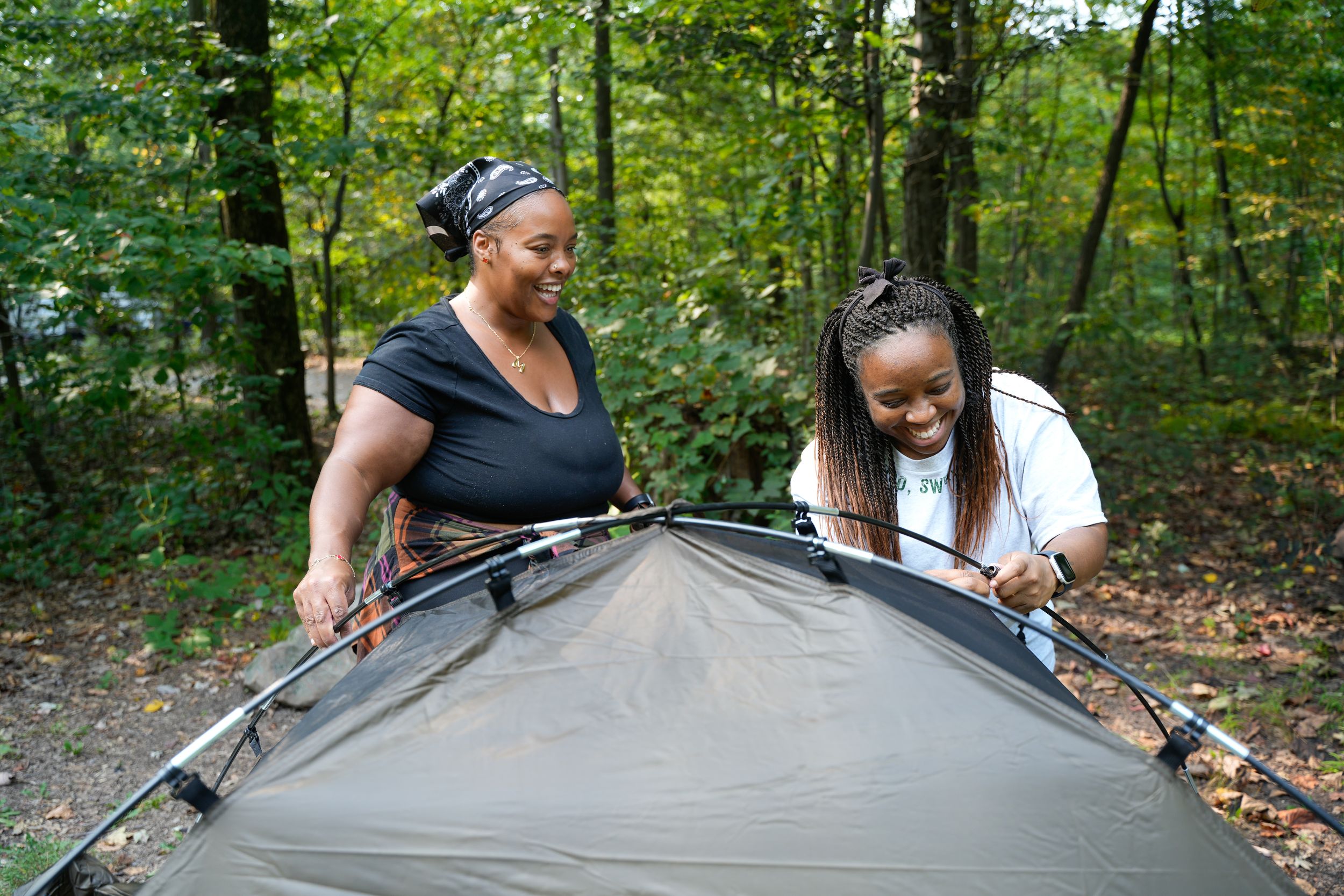 Two women smiling while setting up a tent together in a wooded campsite.