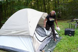 Woman setting up a dome tent at a wooded campsite with a cooler nearby on a cloudy day.