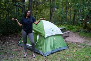 A smiling woman poses proudly next to a green camping tent in a wooded forest campsite.