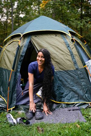 Smiling woman with long braids crouching at the entrance of a green camping tent outdoors.