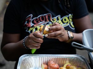 A person peels a potato over a foil pan of vegetables while wearing a black graphic tee.