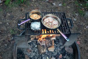 Campfire cooking with pans of food on a metal grill grate over open flames outdoors.