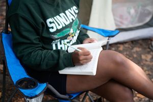A person reads a book in a blue camping chair wearing a green Sundays sweatshirt outdoors.