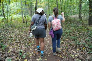 Two women hiking on a forest trail, one carrying a backpack and the other holding a notebook.