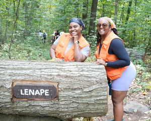 Two women in orange vests smiling behind a large log with a Lenape sign in a forest.