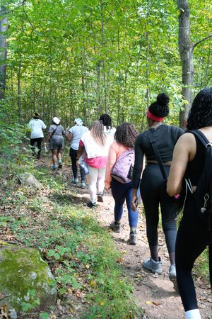 Group of women hiking single file up a rocky forest trail surrounded by green trees.