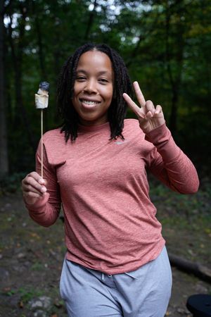 Smiling woman in pink long-sleeve shirt holds roasted marshmallow on stick and flashes peace sign outdoors.
