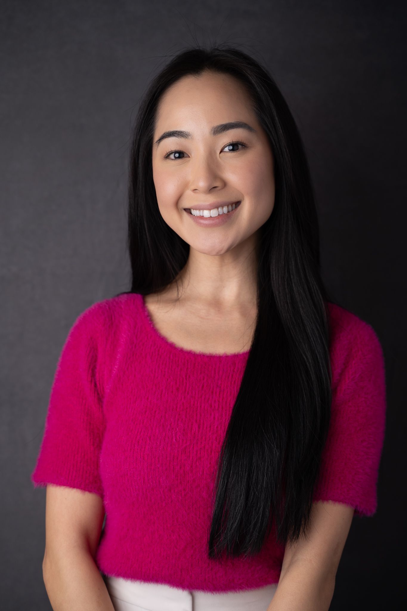 Smiling young Asian woman with long black hair wearing a pink fuzzy sweater against a dark gray background.