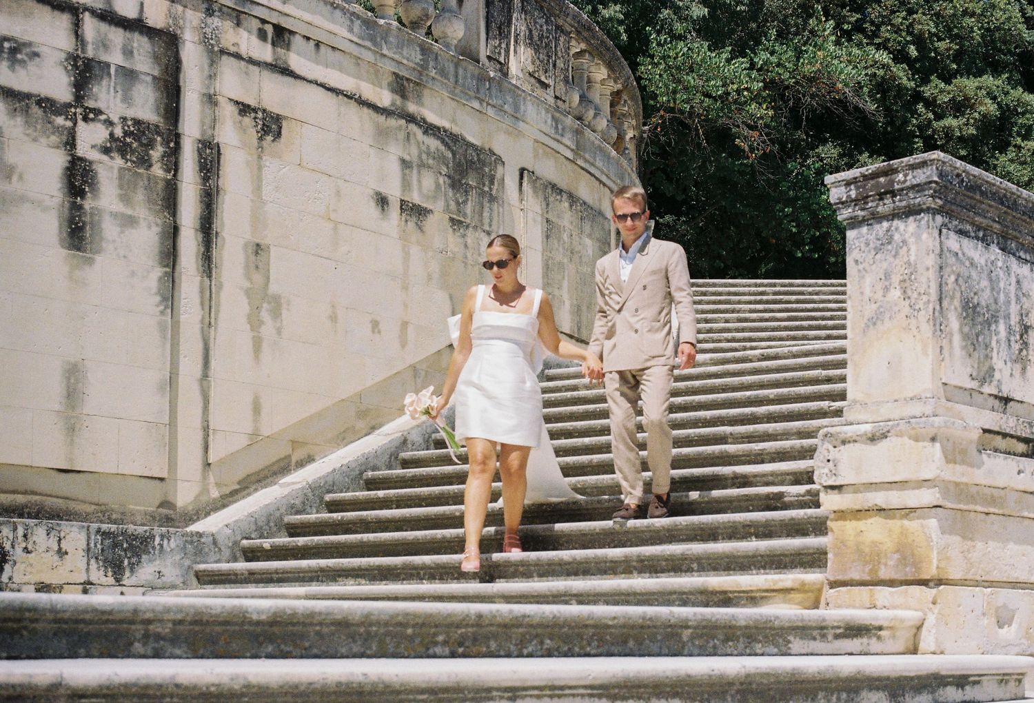 Couple in wedding attire walking down stone steps at an elegant outdoor venue surrounded by lush greenery.