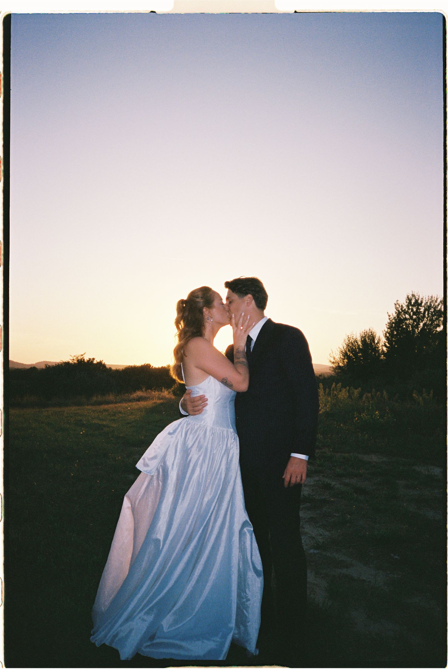 Bride and groom sharing a romantic sunset kiss in an open field, wearing wedding dress and black tuxedo.