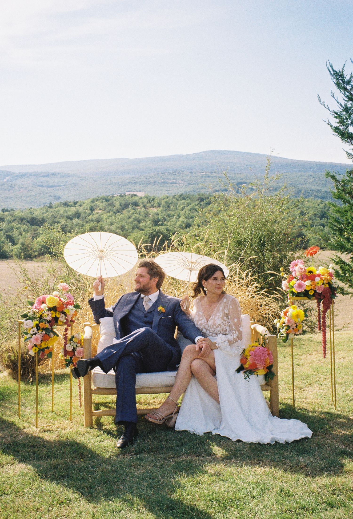 Bride and groom sit outdoors on a sofa with parasols, colorful floral arrangements, and a scenic mountain valley backdrop.