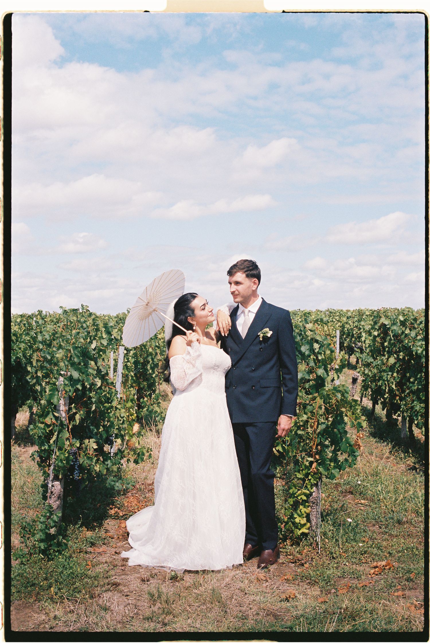 Bride and groom posing in a vineyard, bride holding a white parasol, groom in navy suit on sunny day.