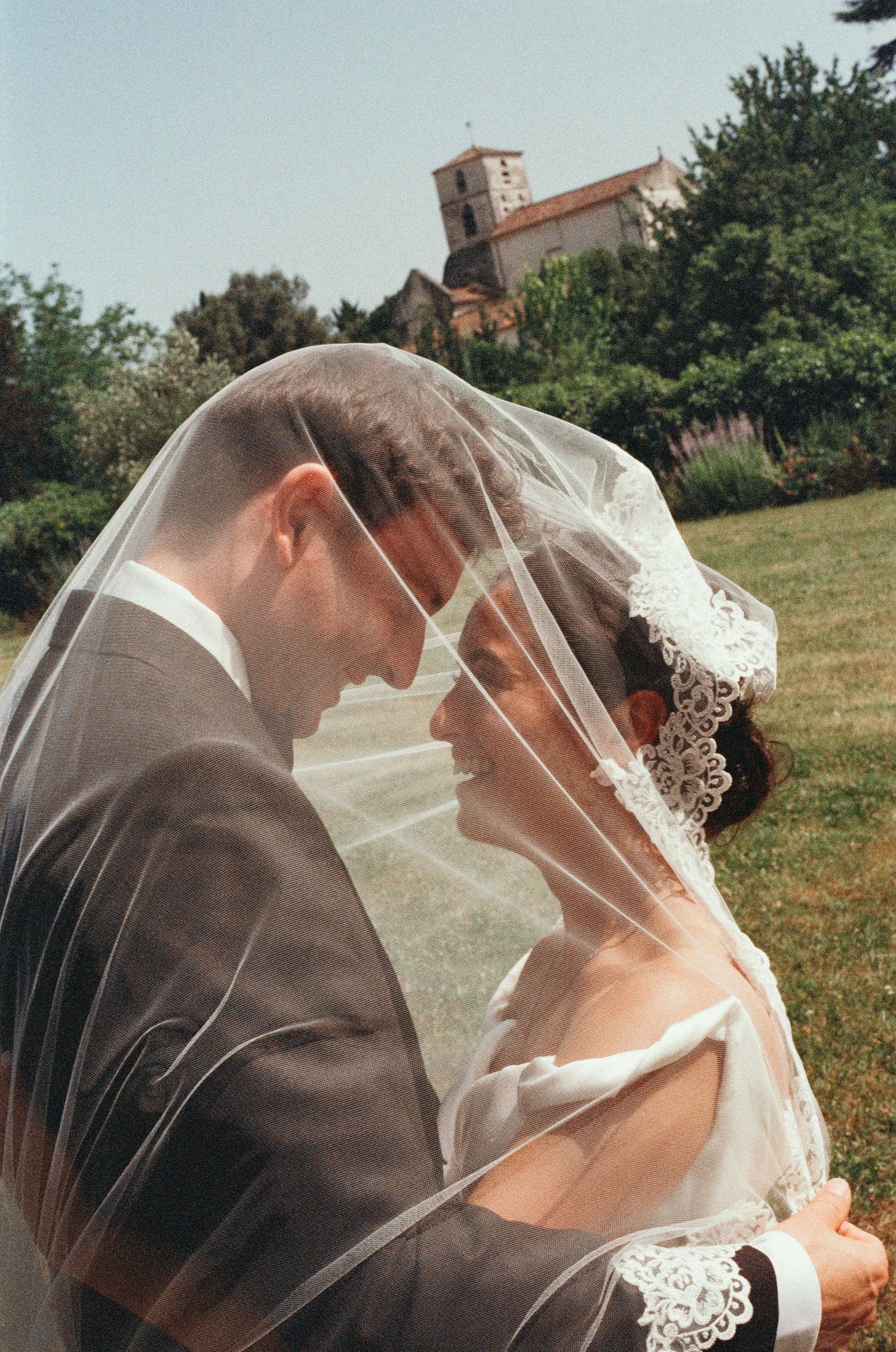 A bride and groom share an intimate moment under a lace wedding veil in a sunlit garden with a church in the background.