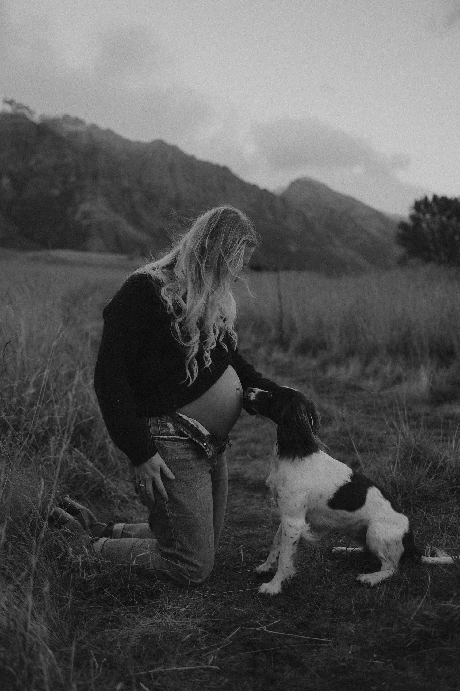 Black and white photo of a woman kneeling in a field petting her Spaniel dog with mountains in the background.
