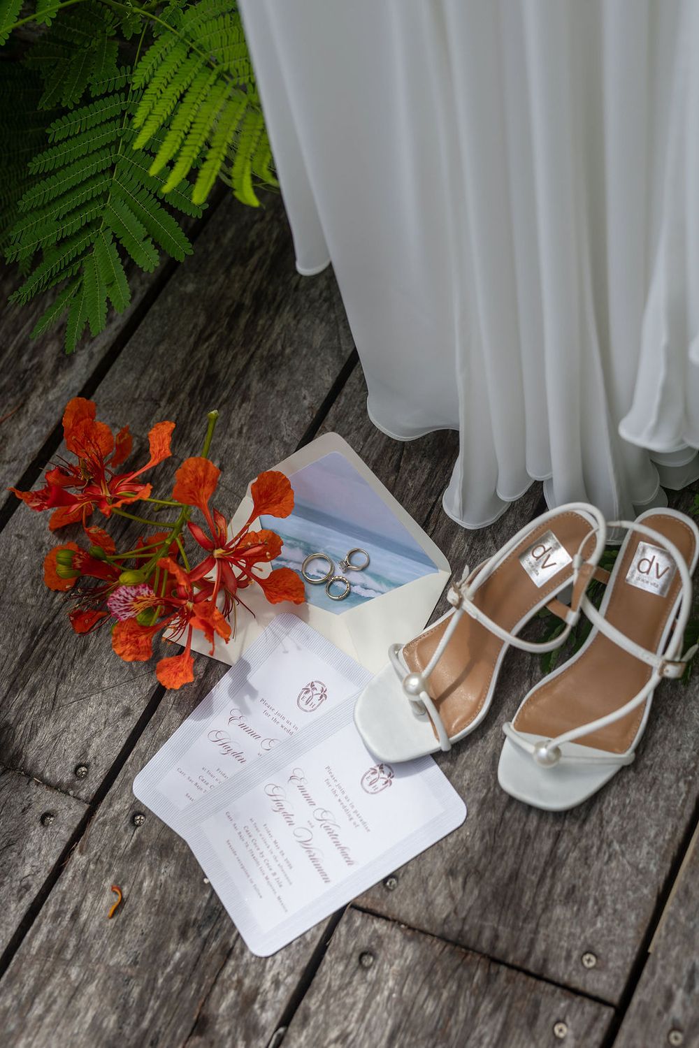 White bridal heels, wedding invitations, red tropical flowers, and rings arranged on rustic wood with a white gown backdrop.